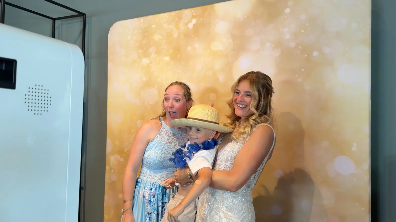 Bruid en gasten met hoed en bloemenkrans poseren voor de photobooth met Gold Lights-achterwand